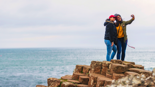 Two visitors standing on the Giant's Causeway's basalt columns near the Middle Causeway, taking a selfie with the ocean in the background under an overcast sky